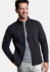 Giacca in pile grigia con colletto alto, zip frontale e tasche laterali. Dettagli di cucitura distintivi sulle spalle; indossata sopra una maglietta grigia chiara.