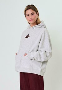 Gaiši pelēks kapuce ar lielu priekšēju kabatu, ribotām aprocēm un brūnu logo uz krūtīm. Mīksts audums un pārspīlēta pielāgošana.