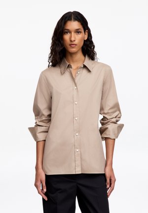 Femme aux longs cheveux bouclés et foncés porte une chemise beige boutonnée avec les manches retroussées et un pantalon noir, debout devant un fond clair uni.