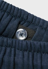 Marineblauer Stoff mit gerippter Textur, ausgestattet mit einem elastischen Bund und einem silbernen Knopf mit Kreuzdesign, festgehalten durch einen schwarzen Riemen.