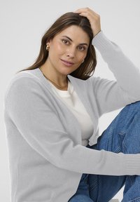 Cardigan grigio chiaro con polsini a coste, design con apertura frontale, indossato sopra una maglietta bianca. La modella è seduta, mostrando una texture morbida e una vestibilità rilassata.