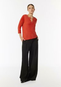 Rote, halbtransparente Bluse mit 3/4-Ärmeln und Reverskragen, kombiniert mit locker sitzenden schwarzen Hosen und spitzen schwarzen Absätzen, minimalistischem Design.