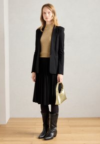 Black blazer over a textured gold sweater, pleated black skirt, black tights, brown cowboy boots, and a metallic gold handbag.