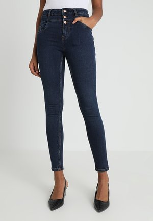 Žena ve tmavě modrých úzkých džínách s vysokým pasem a třemi knoflíky, s černými botami na podpatku s špičatou špičkou, s rukou v kapse.