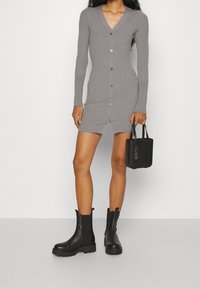 Robe en maille côtelée grise avec manches longues et col en V, ornée de détails à boutons sur le devant. Associée à des bottines noires et un petit sac.
