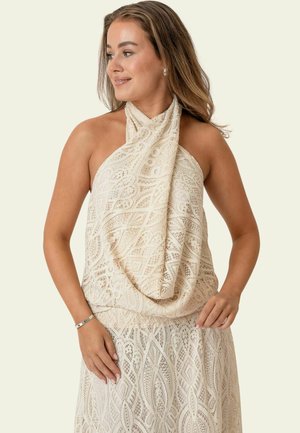 Kobieta w kremowej koronkowej sukience bez rękawów z wiązaniem na szyi, uśmiechająca się i patrząca na bok na neutralnym tle.