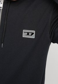 Felpa nera con zip realizzata in tessuto testurizzato, presenta un'etichetta con logo argentato e accenti di cuciture visibili. Design liscio e aderente.