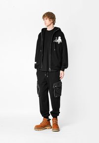 Joven con sudadera negra con estampado gráfico, pantalones cargo negros con grandes bolsillos y botas de cordones marrones, de pie frente a un fondo blanco.