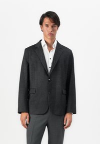 Junger Mann in einem dunkelgrauen Hahnentritt-Blazer über einem weißen Hemd und grauen Hosen, der vor einem schlichten weißen Hintergrund steht.