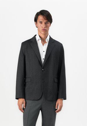 Junger Mann in einem dunkelgrauen Hahnentritt-Blazer über einem weißen Hemd und grauen Hosen, der vor einem schlichten weißen Hintergrund steht.