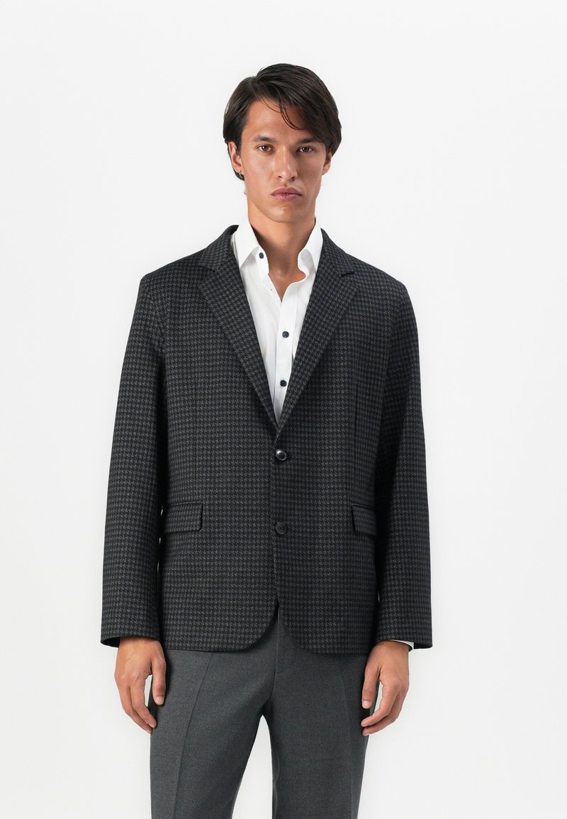 Junger Mann in einem dunkelgrauen Hahnentritt-Blazer über einem weißen Hemd und grauen Hosen, der vor einem schlichten weißen Hintergrund steht.