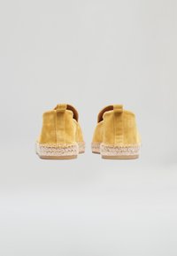 Para żółtych zamszowych espadryli widzianych od tyłu, z plecionymi podeszwami z juty i pętelkami do zakładania, na jasnym tle.