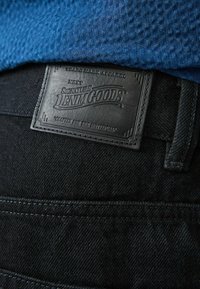 Svarta jeans i denim med en texturerad blå skjorta delvis synlig ovanför. Jeansens har en läderpatch med präglad text.