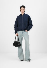 Marineblå bomberjakke med glidelås foran og snøring i nederkant, sammen med lyseblå utsvingte jeans og en liten svart håndveske.
