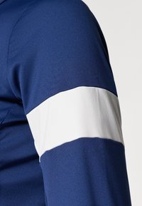 Camisa azul marinho com uma faixa branca canelada em torno da manga esquerda. O tecido é suave, com um ligeiro brilho e um design ajustado.