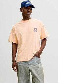 Bărbat tânăr purtând un tricou supradimensionat de culoare piersică deschisă cu un logo mic, blugi albastru deschis și o șapcă de baseball albastră, stând în fața unui fundal simplu.