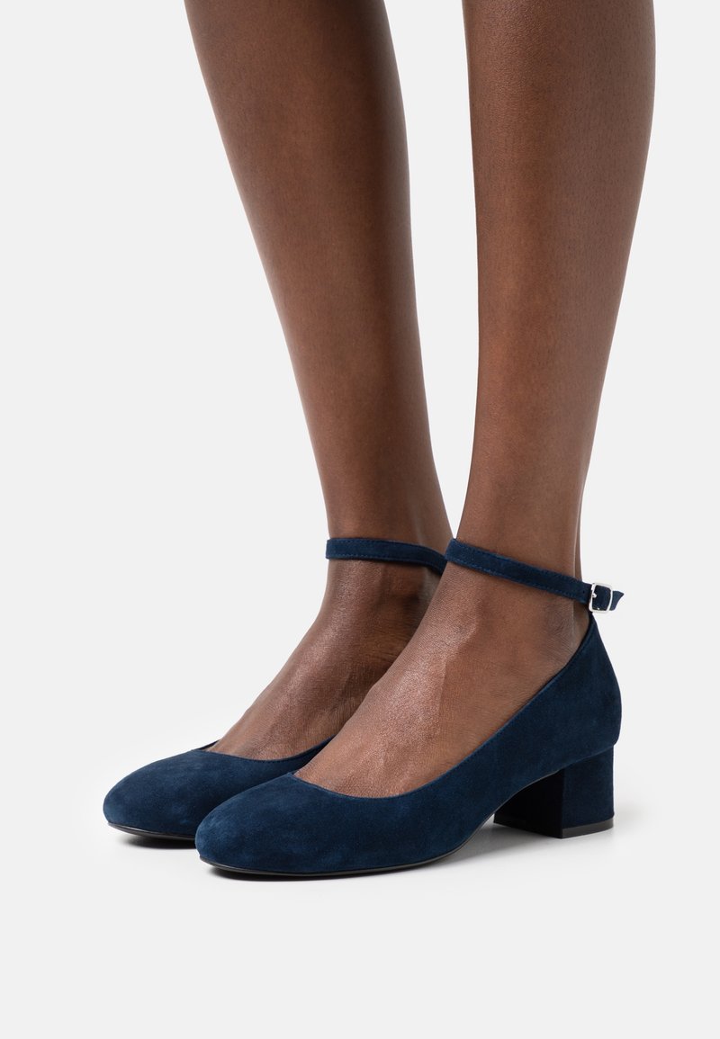 Jambes à peau foncée portant des chaussures à talons blocs en daim bleu marine avec brides à la cheville et boucles argentées sur un fond clair uni.
