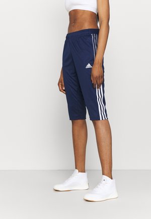 Vrouw die marineblauwe Adidas atletische shorts tot op de knie draagt met witte strepen, een witte sportbeha en witte hoge sneakers, zijaanzicht.