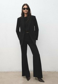 Blazer negro a medida con solapas marcadas, pantalones a juego de talle alto y tacones puntiagudos. Gafas de sol completan el look monocromático.