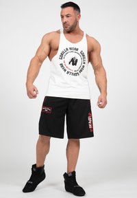 Wit mouwloos tanktop met een zwarte grafische print, gecombineerd met zwarte shorts met rode accenten en een logo. Zwarte sportschoenen.