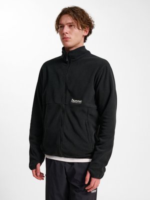 Junger Mann trägt eine schwarze Fleecejacke mit Reißverschluss und passende schwarze Hose vor weißem Hintergrund.