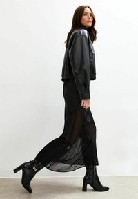 Veste en cuir noir cropped, robe maxi noire transparente avec des fentes latérales, et bottes noires à talons hauts jusqu'aux genoux avec bouts carrés et détails.