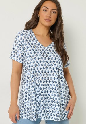 Femme aux longs cheveux bruns portant un haut blanc à manches courtes orné de petits motifs floraux bleus, associé à un pantalon bleu clair.