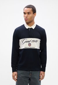 Polo cu mânecă lungă de culoare bleumarin, cu dungi orizontale crem, având inscripția "Gant 1949" și un logo. Amestec de bumbac, cu guler standard.
