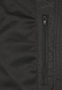 Chaqueta negra con un panel de malla texturizada en un lado y tela lisa en el otro, con un bolsillo con cremallera para almacenamiento.