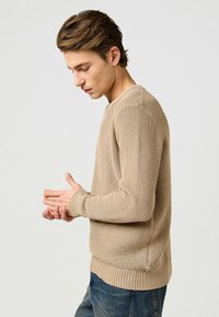 Beiger Strickpullover mit strukturiertem Muster, rundem Halsauschnitt sowie geripptem Saum und Bündchen, getragen von einem Model mit gefalteten Händen.