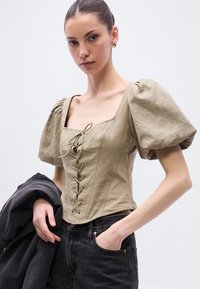 Χακί μπλούζα με φουσκωτά μανίκια, τετράγωνη λαιμόκοψη, δέσιμο στο μπροστινό μέρος και ύφασμα από μείγμα βαμβακιού και λίνου. Συνδυασμένη με ψηλόμεσα σκοτεινά τζιν.