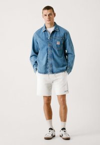 Chaqueta de mezclilla color azul claro con bolsillo frontal, camiseta blanca, pantalones cortos blancos y zapatillas de deporte en blanco y negro con detalle de textura. El modelo está de pie en posición recta.