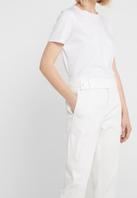 Kobieta w prostym, białym t-shircie z krótkim rękawem, zapiętym w wysokie białe spodnie z paskiem w talii i kieszeniami, stojąca na tle białym.