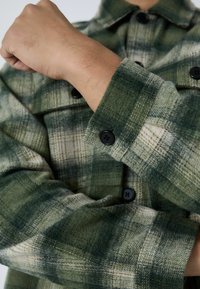 Zaļš un krēma krāsas krekliņš ar platu rakstu, ar pogām uz aproces, kam ir teksturēts apdare un divas melnas pogas uz piedurknes.