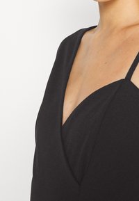 Femme portant une robe noire sans manches à col en V avec fines bretelles, montrant le décolleté et le haut de la poitrine.