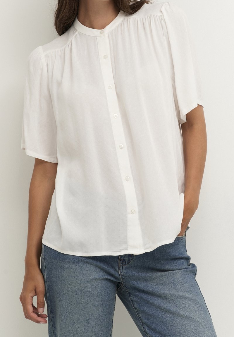 Femme portant un chemisier blanc à manches courtes boutonné avec de légers plis aux épaules et un jean bleu, main reposant dans la poche.