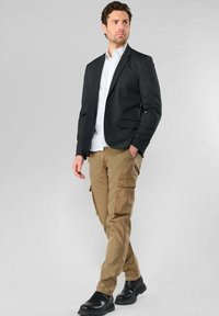 Blazer gris foncé sur une chemise blanche, associé à un pantalon cargo beige. Chaussures noires et poches discrètes sur le pantalon visibles. Aspect propre et bien ajusté.