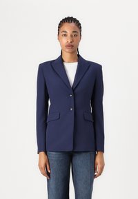 Marineblaues Blazer aus glattem Stoff, mit einem Reverskragen, zwei Fronttaschen und zwei Knöpfen. Getragen über einem weißen Oberteil.
