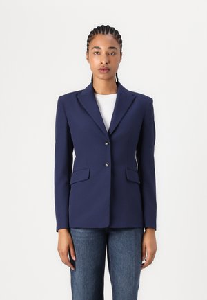 Navy blue blazer made of smooth fabric, featuring a notched lapel, two front pockets, and two buttons. Worn over a white top.