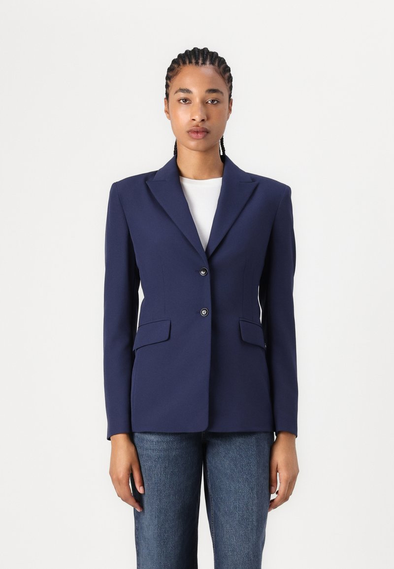 Blazer blu navy realizzato in tessuto liscio, con rever a punta, due tasche frontali e due bottoni. Indossato sopra un top bianco.
