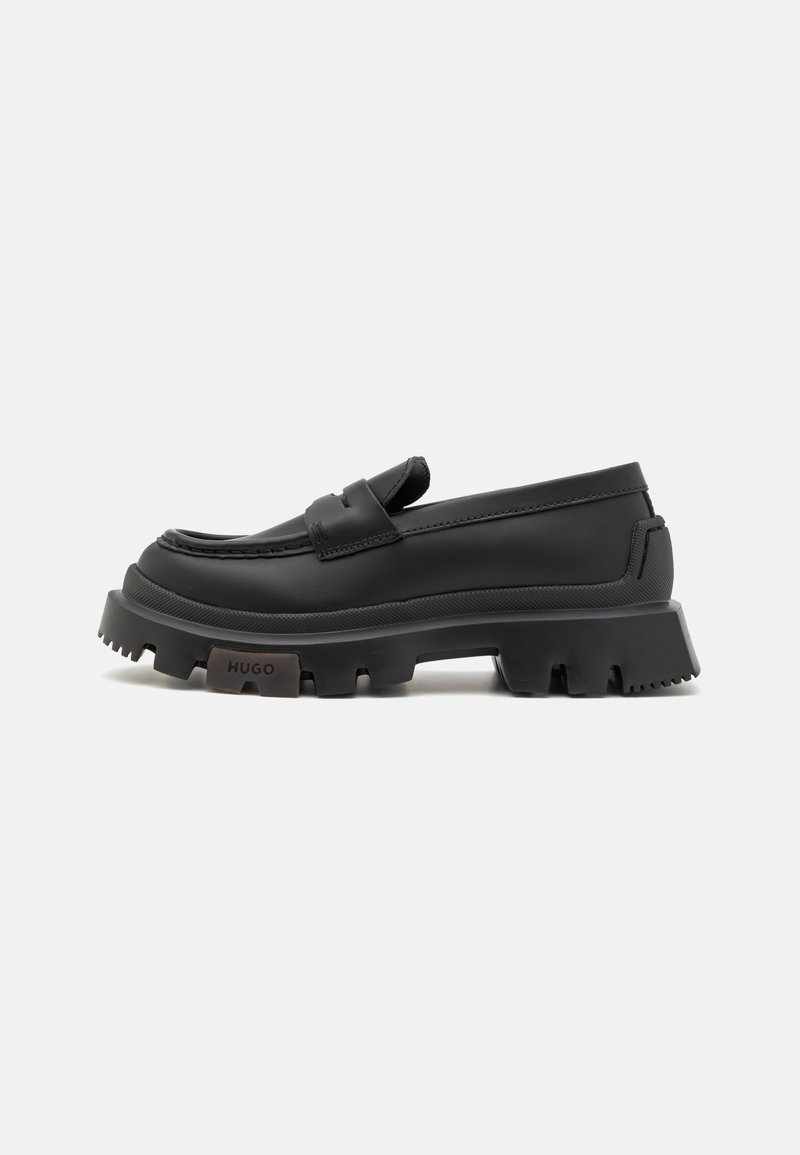 Mocassin en cuir noir à semelle épaisse, avec un détail de bride et le logo « HUGO » sur la semelle, présenté sur un fond blanc uni.