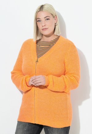 Oranžový pletený kardigan se zipem, designem do V a přírodní strukturou. Nosený přes černé a oranžové pruhované tričko, spárovaný s tmavými džíny.