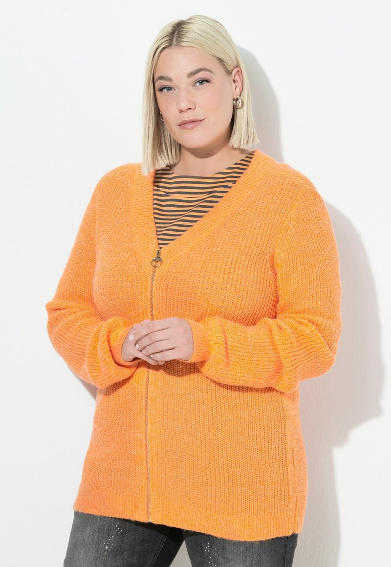 Cardigan di maglia arancione con chiusura a zip, design a scollo a V e texture a coste. Indossato sopra una maglietta a righe nera e arancione, abbinato a jeans scuri.