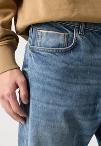 Vaqueros azules deslavados con corte recto, que presentan costuras naranjas, cinco bolsillos y una tela texturizada. Una mano reposa en el costado.