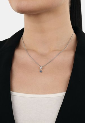 Vrouw draagt een zilveren ketting met een kleine ronde blauwe edelsteen hanger, gekleed in een zwarte blazer en een witte top.