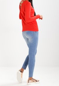 Rode langwerpige top met een v-achterkant, gecombineerd met lichtblauwe skinny jeans en metallic roségouden instap schoenen. Soepele stof en pasvorm.