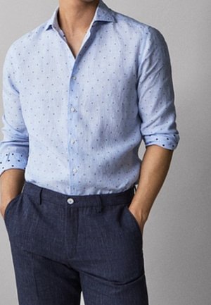 Homme portant une chemise à boutons bleu clair à motif, manches retroussées, et un pantalon bleu foncé, les mains dans les poches.