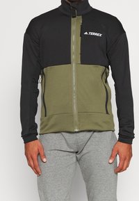Kurtka zip-up Terrex z czarną górną częścią i oliwkowym dolnym fragmentem, wyposażona w boczne kieszenie i wysoki kołnierz dla dodatkowego ciepła.