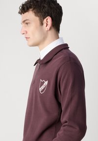 Burgundijska majica s patentnim zatvaračem, koja ima rebrasti ovratnik, iz vezenu oznaku na prsima i mekan, teksturiran materijal.