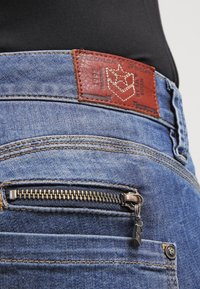 Modré džínové kalhoty s hnědým koženým štítkem, viditelnými švy, kapsou na zip a tmavým pasem.